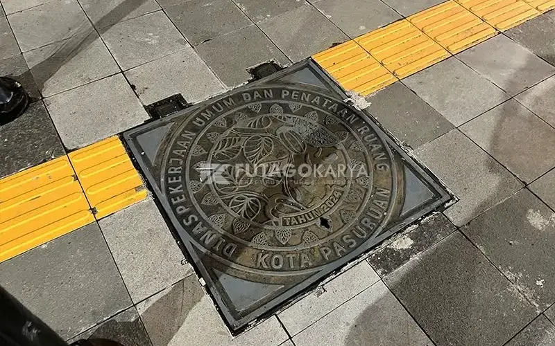 Manhole cover besi cor berbentuk bulat terpasang pada jalur pedestrian dengan guiding block kuning
