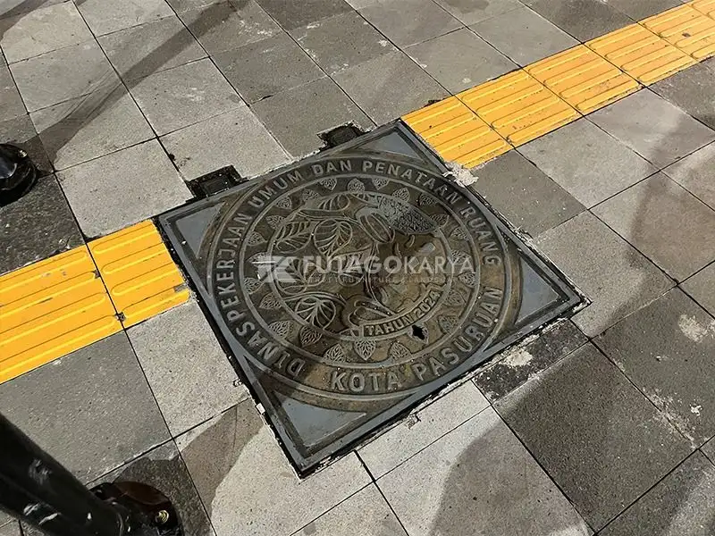 Manhole cover besi cor berbentuk bulat terpasang pada jalur pedestrian dengan guiding block kuning